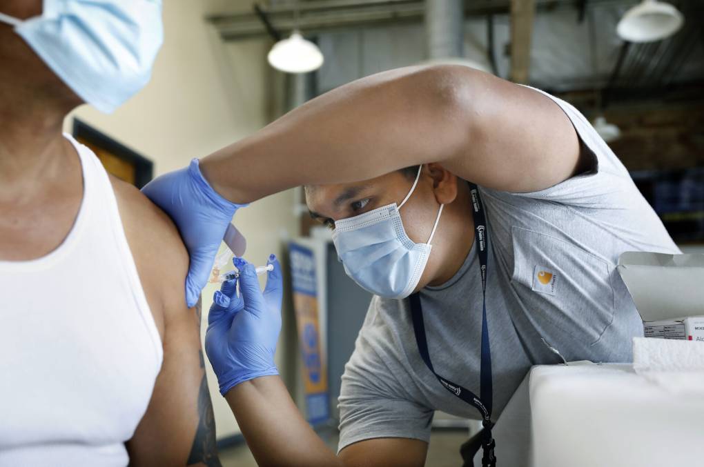 A person wearing a mask and latex gloves administers a shot in another person's arm.