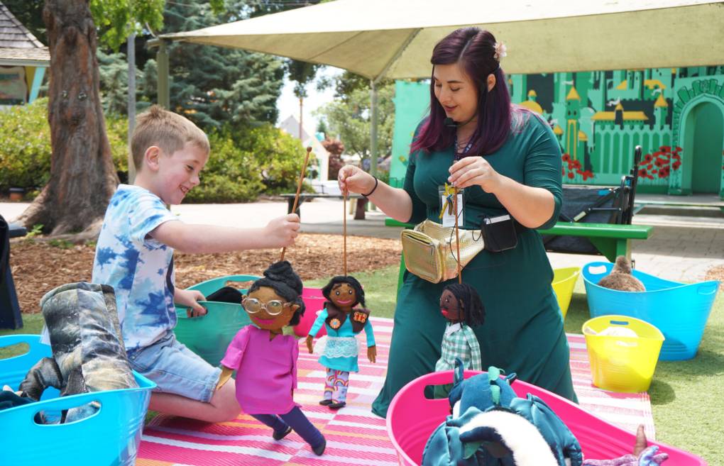 A woman wearing a green dress holds puppets in front of a young boy wearing a blue shirt.