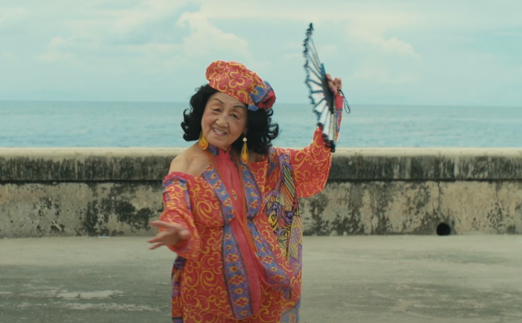 A senior Asian woman with black hair, wearing a flowing gown and matching hat, dances with a fan near the Pacific Ocean.